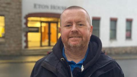 The photo shows a man with a ginger beard and very short hair. He is wearing a navy blue jumper and coat and smiling at the camera. He is standing outside, and behind him is a brick building.
