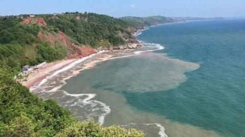 A picture of Oddicombe Beach in Torquay from above. The cliffs curve to the left of the image with waters lapping onto a curve of sand below. There is red rockface and green vegetation covered cliffs.