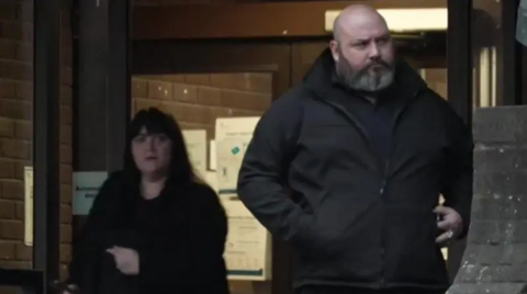 A picture of Hayley Bell, on the left, and Richard Elkin, on the right, coming out of Portsmouth Crown Court. Hayley Bell is wearing a black coat and has long, dark hair. Richard Elkin is bald and has a white and grey beard.