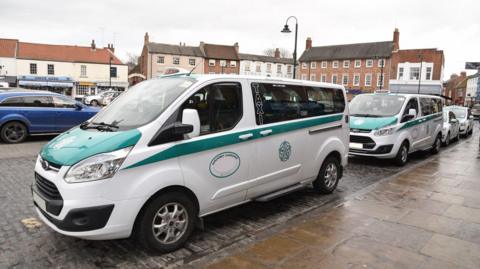 A line of taxis at a taxi rank on a cobbled, wet road in the middle of a town. The vehicles are white with turquoise markings. Each vehicle has an oval marking on its door and a "taxi" light on the roof. The sky is overcast.