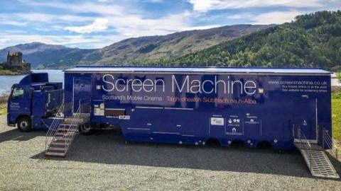 A navy blue Screen Machine lorry is parked next to Loch Duich with Eilean Donan Castle in the background. The sky is bright with a few clouds and forests can be seen on the hillside on the other side of the loch.