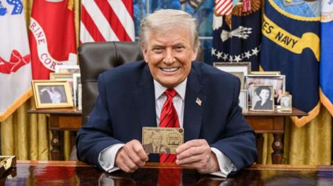 President Donald Trump smiles for the camera in the White House and he holds up a Trump Gold Card