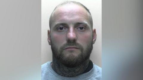 A mugshot of Liam Smith. He has short light brown hair and a brown beard and moustache. He has brown eyes and his neck has blue tattoos. He is wearing a light grey sweatshirt.