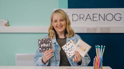 Anwen is looking at the camera, sitting at a table holding some of her cards up in both hands and smiling. she is wearing a light jean jacket. She has should length blonde hair. Her company logo the name Draenog is on a wall in the background