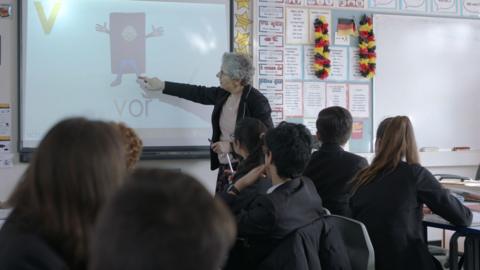 A teacher in a classroom, pointing at whiteboard.