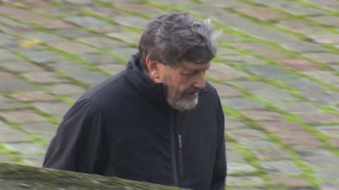 Alan Harris walks towards Winchester Crown Court. He is wearing a black coat which has been zipped up. He has short greying dark hair and a greying beard.