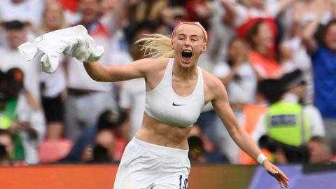 Chloe running towards the camera with her arms to her sides, wearing white shorts and white sports bra with her England shirt twirled up in her right hand, screaming with joy, blonde hair flying