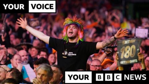 A crowd of people watching a darts contest at Glasgow's Hydro. A man in the middle of the crowd is on his feet, wearing a multi-coloured wig and a black t-shirt. His arms are out-stretched and his mouth open wide. He's enjoying himself.