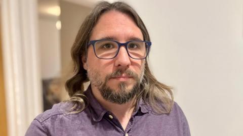 A man in a purple collared shirt with long hair and glasses. He has a stern expression.