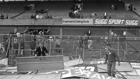 A black and white image looking up into a football stand with people stood around in the aftermath of the disaster. There is debris lying around, including advertising boards which were used as stretchers. A few men try and move the boards.