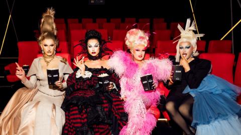 Silllexa Diction wears a golden gown, Bones is in a black and red stripped outfit, Elle Vosque is in a pink fluffy outfit and Catrin Feelings wears a black blazer and blue tulle skirt.