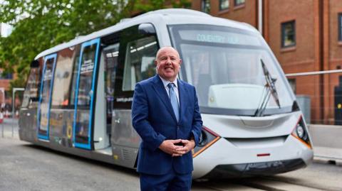 Jim O'Boyle stands in front of a silver tram vehicle. He is wearing a blue suit and blue tie.