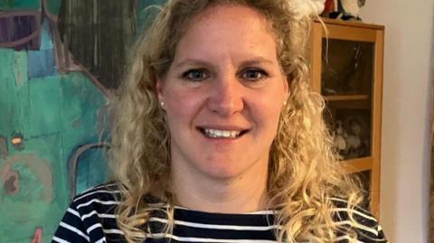 Camilla Notley has curly blonde hair and is smiling. She is standing in front of one of Malcolm Race's abstract paintings. Teal and turquoise shapes with grey lines.