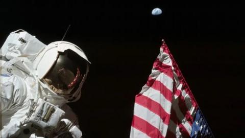 Harrison Schmitt is photographed next to the United States flag on the lunar surface during the Apollo 17 mission. The highest part of the flag appears to point toward our planet Earth in the distant background. Its red and white stripes are also reflected in the visor of Schmitt's helmet.