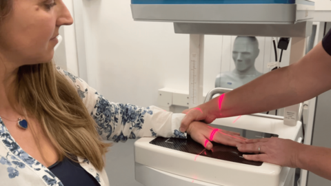 A woman sits with her left hand on a 3D imaging device. There is a bright red cross on her hand created by a light on the device.