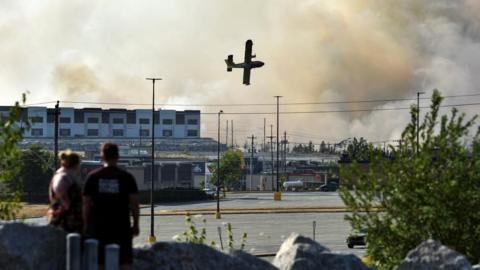 A man and a woman watch a water bomber try to extinguish a wildfire near Bayers Lake at the edge of Halifax, Nova Scotia. Industrial buildings are seen in the background, along with clouds of smoke and the plane.
