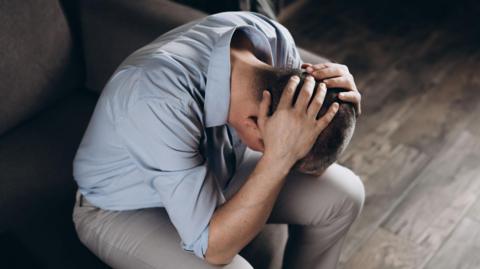 A man in despair with his head in his hands. He is wearing a blue shirt and light trousers. We cannot see his face but he has short, cropped dark hair.