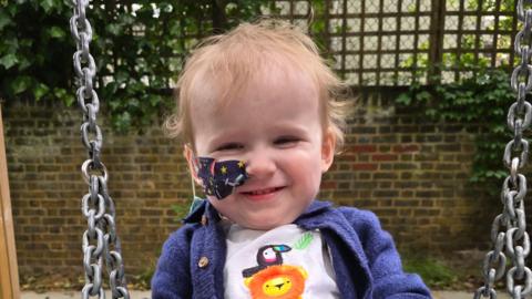 A young boy smiling at the camera wearing a lion and parrot T-shirt and blue jacket. He is sitting on a swing outside. He has a space-themed plaster on his cheek which is holding a tube in place in his nose.