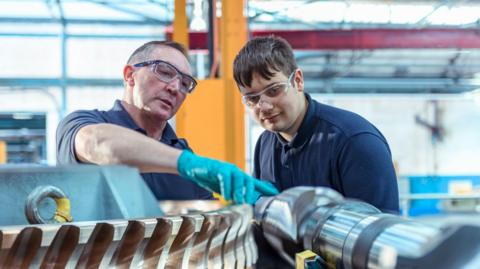 An older man wearing blue plastic gloves is showing a large piece of metal equipment to a young man, both of whom are wearing short-sleeved navy polo shirts. They also have protective eye wear on and appear to be stood in a large factory warehouse, which is blurred in the background behind them.