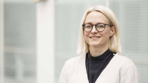 A blonde woman with glasses looks at the camera and smiles. She is wearing a black top and a white cardigan, and is standing in front of a grey background.
