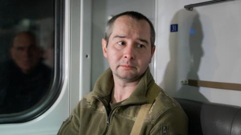 Denys, sitting on a train, looks down away from the camera, lips downturned. He is wearing military fatigues.