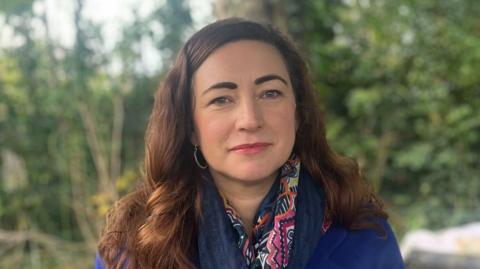 Margaret Gibney sitting outdoors, looking at the camera. She has long, dark, wavy hair and is wearing a long royal blue coat with a navy and pink scarf and hooped earrings. There are trees in the background.