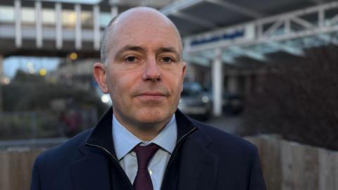 Roland Sinker looking into the camera outside the front of Addenbrooke's Hospital. He is wearing a navy fleece over a blue shirt with a purple tie.