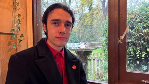 A man wearing a black jacket and a red shirt, wearing a badge that says 'Labour'. He is sat down inside a cafe, with a window behind him showing a green lawn.