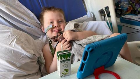 Connor sitting up in a hospital bed and cuddling a grey fluffy blanket. He is holding a Maui toy from Disney's Moana movie, and watching something on a blue iPad propped up in front of him, next to a carton of apple juice. He is smiling at the camera with a feeding tube in his right nostril, secured to his cheek with a large blue plaster.