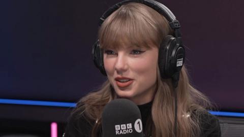 Taylor Swift wearing black headphones and in front of a BBC Radio 1 microphone