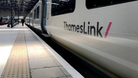 A ThamesLink train is stationary at a train station platform. The train is white with blue doors and has the ThamesLink writing and logo on the side.