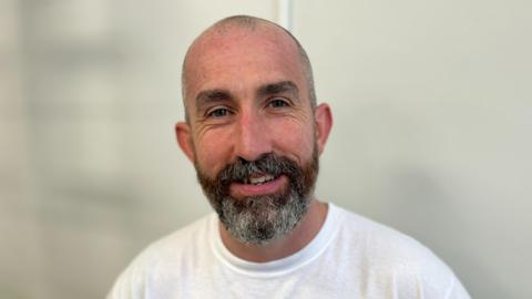 Gerard Donaghy is standing in front of a white wall, wearing a white t-shirt and is smiling at the camera. He has a bald head and a dark beard with grey hair through it.