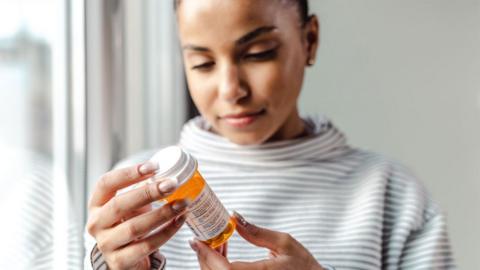 A young women with a stripey top holds a bottle of pills and reads the label.