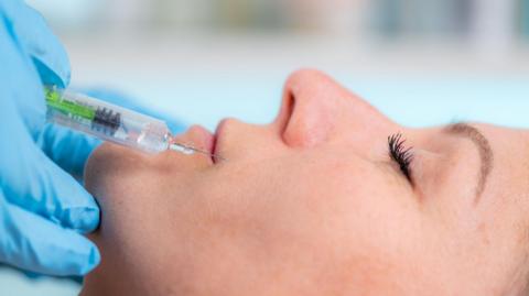 A close-up of a woman's face, side on, getting an injection of filler into the skin surrounding her lips. The needle hovers above her lip, held by a hand wearing blue gloves.