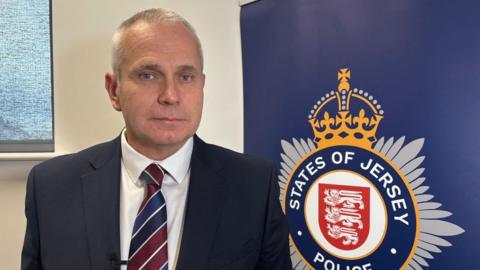 A man in a blue suit and white top and blue and red tie. He is stood in front of a States of Jersey Police sign. The wall behind him is cream.