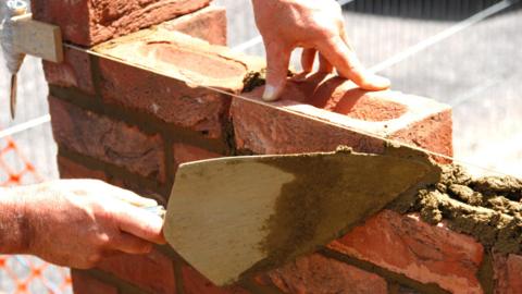 A person building a brick wall with red brick and cement holding a trowel.
