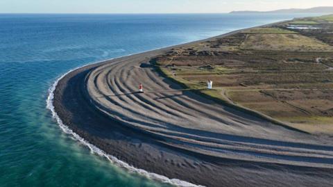 An ariel view of the Point of Ayre in the north of the Isle of Man, you can see the turqoies water hitting the arching headland, there is a lighthouse on the beach.