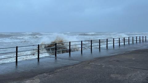 The Irish Sea seen from the promenade in Maryport. Tall waves are crashing on the promenade and the sky is grey with heavy clouds.