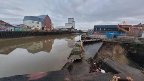 The photo shows the rather industrial setting of the River Hull, with concrete steps and discarded rubbish. An old rusty and broken foot bridge is covered by moss. The water is dirty and brown.