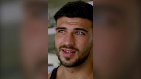 Tommy Fury looks away from the camera. He has short brown hair, blue eyes and wears a black vest.