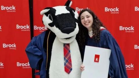 Eileen standing next to a badger mascot. She's in a graduation gown and is standing in front of a red background with the Brock University logo printed on it.