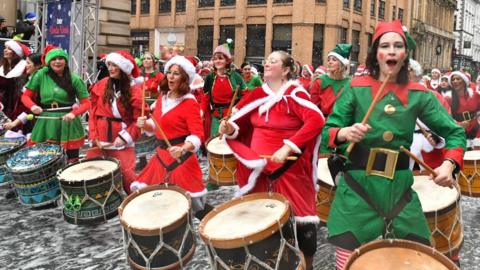 Rows of women in red santa suits and green elf suits sing and play traditional drums with their drumsticks. Old buildings rise up behind.