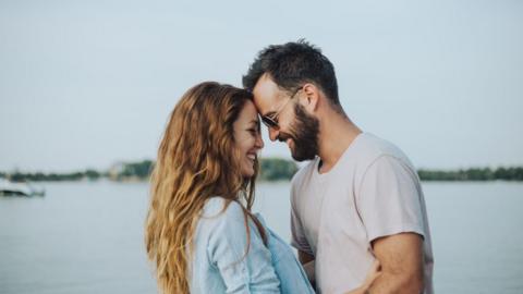 A man and a woman smile as they stand facing each other in front of a water body