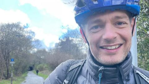 Euan is wearing a blue cycling helmet. He is smiling at the camera. The Roseburn Path is in the background.