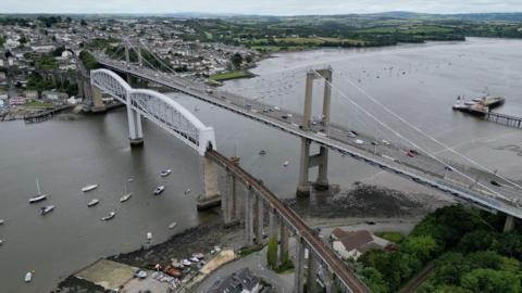 The photo shows an aerial view of the Tamar Bridge, a suspension bridge over the River Tamar. Next to it is a rail crossing. Houses and green fields are seen in the distance.