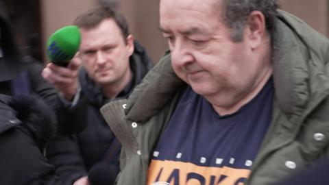 Gerard Woodhouse, who is wearing an olive green overcoat and a Jack & Jones t-shirt, is seen leaving court. He has his head down and is surrounded by reporters, some with microphones.
