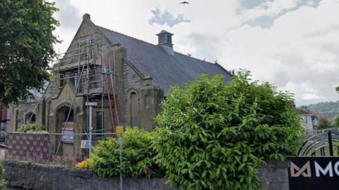 A church with scaffolding on the exterior. It is set back from the street and surrounded by a low, stone wall and shrubbery.