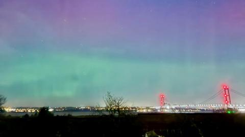 Humber Bridge lit up at night with red and white lights and the sky is purple and blue. There is a river and trees.