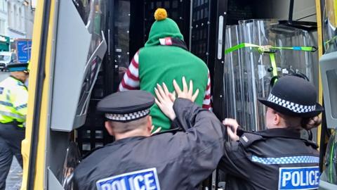 Police officers push a man wearing a green elf costume with a small hat and a yellow bobble.