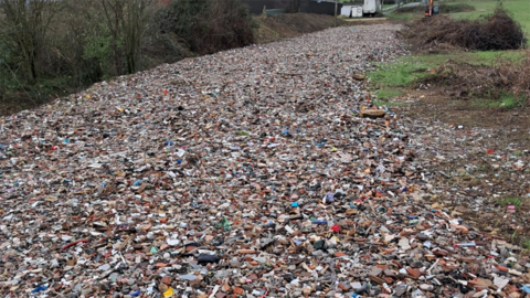 A field that is completely covered in rubble and mixed waste, including broken bricks, stones and bits of plastic. Bushes and fields line the sides.